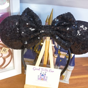 May include: Black sequin Minnie Mouse ears headband with a large black bow. The headband is on a wooden stand with a sign that reads "Good To Be Ear".