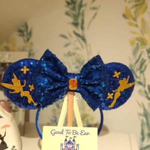 May include: Blue and gold sparkly Minnie Mouse ears with a Tinkerbell design. The ears are on a stand with a sign that reads "Good To Be Ear" and "Designs From Ear To There".