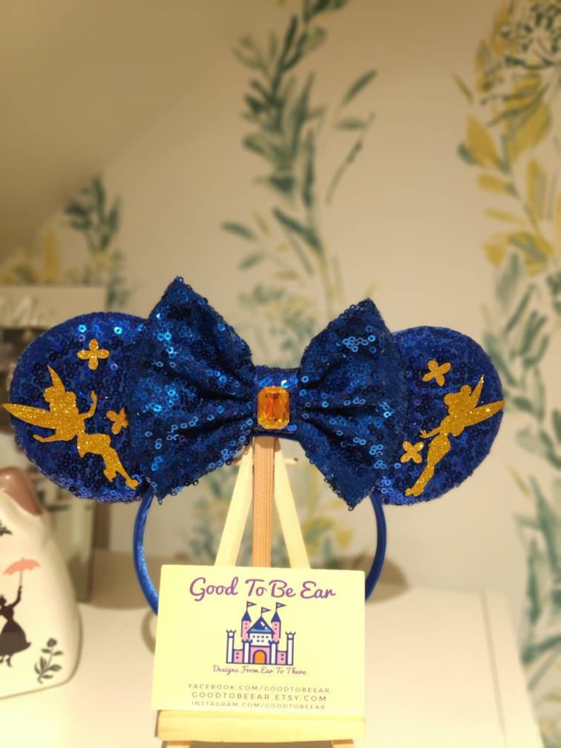 May include: Blue and gold sparkly Minnie Mouse ears with a Tinkerbell design. The ears are on a stand with a sign that reads "Good To Be Ear" and "Designs From Ear To There".