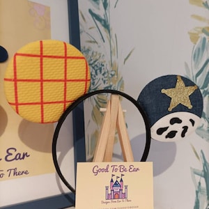 Op de afbeelding: Een paar handgemaakte Mickey Mouse oren met een geel en rood geruit patroon op het ene oor en een blauw denim oor met een gouden ster en witte koe vlekken op het andere. De oren worden getoond op een klein houten ezel met een visitekaartje waarop staat "Good To Be Ear Designs From Ear To There" met contactgegevens voor Facebook, Etsy en Instagram.