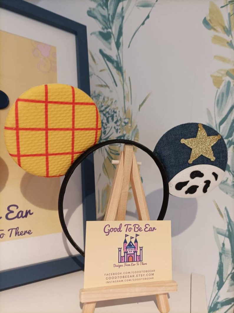 Op de afbeelding: Een paar handgemaakte Mickey Mouse oren met een geel en rood geruit patroon op het ene oor en een blauw denim oor met een gouden ster en witte koe vlekken op het andere. De oren worden getoond op een klein houten ezel met een visitekaartje waarop staat "Good To Be Ear Designs From Ear To There" met contactgegevens voor Facebook, Etsy en Instagram.