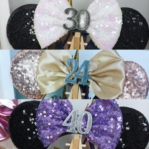 May include: Three pairs of black and sparkly mouse ears with a satin bow and a silver number on top. The numbers are 30, 24, and 40.