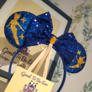 May include: Blue and gold sequined Minnie Mouse ears with a Tinkerbell design. The ears are on a headband and have a bow in the center.