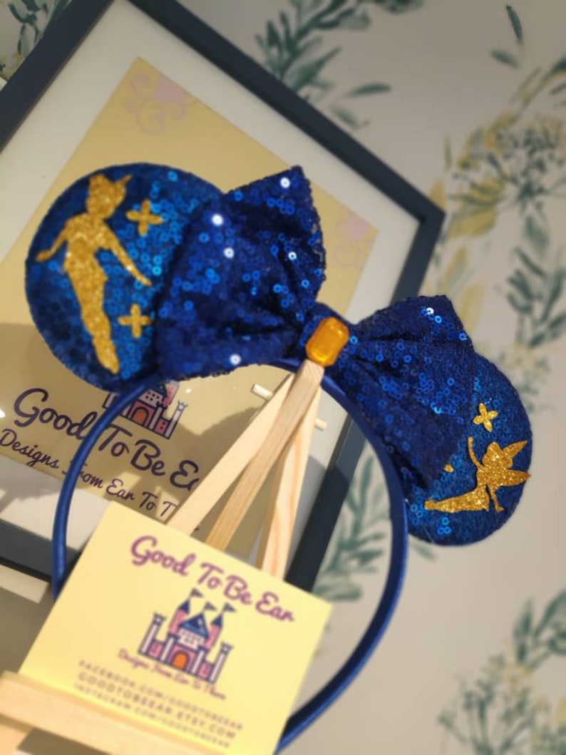 May include: Blue and gold sequined Minnie Mouse ears with a Tinkerbell design. The ears are on a headband and have a bow in the center.