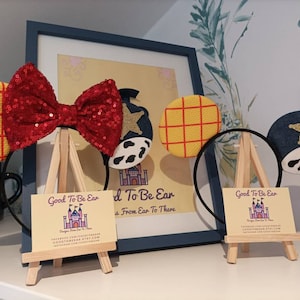 Op de afbeelding: Twee paar handgemaakte Disney-muisoren op een display. Het ene paar heeft een rode pailletten strik met een geel en rood geruit oor en een blauw oor met een witte ster en een koe-print. Het andere paar heeft een geel en rood geruit oor en een blauw oor met een witte ster en een koe-print. Beide paren worden getoond op kleine houten ezels met visitekaartjes waarop "Good To Be Ear" en "Designs From Ear To There" staat.