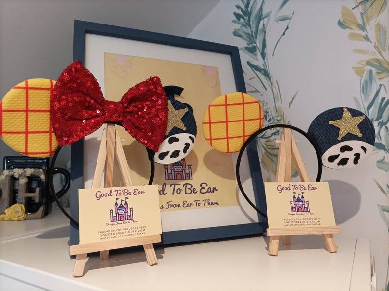 Op de afbeelding: Twee paar handgemaakte Disney-muisoren op een display. Het ene paar heeft een rode pailletten strik met een geel en rood geruit oor en een blauw oor met een witte ster en een koe-print. Het andere paar heeft een geel en rood geruit oor en een blauw oor met een witte ster en een koe-print. Beide paren worden getoond op kleine houten ezels met visitekaartjes waarop "Good To Be Ear" en "Designs From Ear To There" staat.