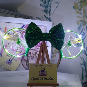 May include: A pair of green sequined headband ears featuring two circular frames with integrated LED lights. Each frame contains a white fairy silhouette with yellow wings. A large green sequined bow sits in the centre. A small sign reads Designs From Ear To There Good To Be Ear.