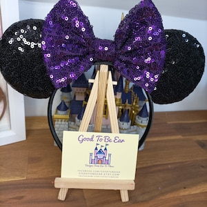 May include: A black headband with sequined mouse ears and a large purple sequined bow. A small wooden easel displays a business card with the text "Good To Be Ear" and a castle illustration.