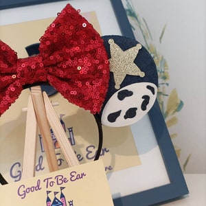 Op de afbeelding: Een rood, met pailletten bezet haarband met een blauw en wit koe-print oor en een gouden ster op het andere oor. Het haarband wordt getoond op een houten ezel met een visitekaartje waarop "Good To Be Ear Designs From Ear To There" staat.