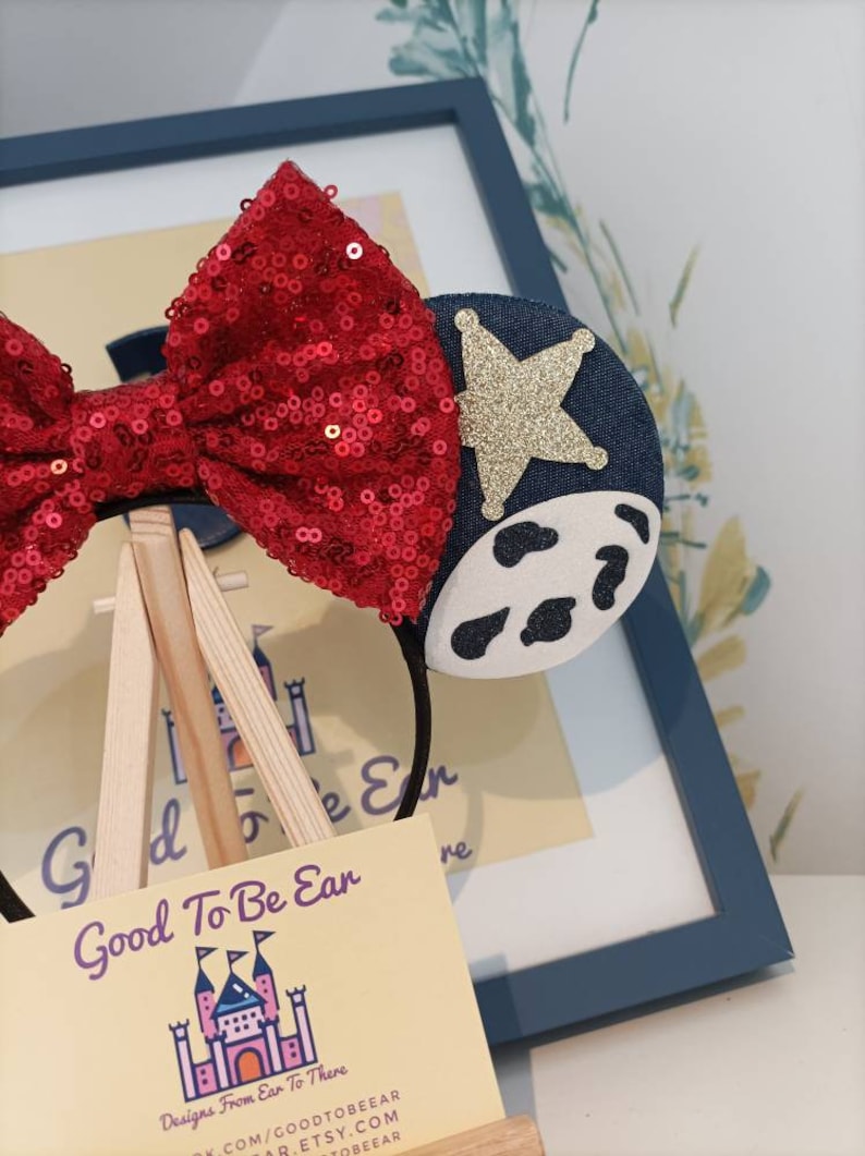 Op de afbeelding: Een rood, met pailletten bezet haarband met een blauw en wit koe-print oor en een gouden ster op het andere oor. Het haarband wordt getoond op een houten ezel met een visitekaartje waarop "Good To Be Ear Designs From Ear To There" staat.