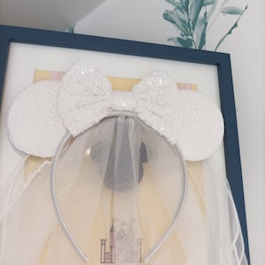 May include: White sequined Minnie Mouse ears with a white tulle veil attached to a silver headband. The ears have a large bow in the centre.