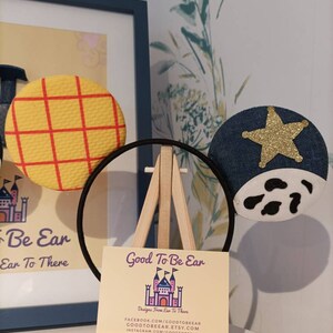 Op de afbeelding: Een paar handgemaakte Mickey Mouse oren met een geel en rood geruit stoffen oor aan de linkerkant en een blauw denim oor met een gouden ster en een wit koe-print aan de rechterkant. De oren zitten op een zwarte hoofdband en worden getoond op een klein houten ezel met een visitekaartje waarop staat "Good To Be Ear Designs From Ear To There" met contactgegevens voor Facebook, Etsy en Instagram.