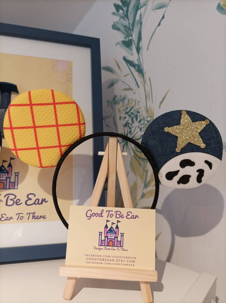 Op de afbeelding: Een paar handgemaakte Mickey Mouse oren met een geel en rood geruit stoffen oor aan de linkerkant en een blauw denim oor met een gouden ster en een wit koe-print aan de rechterkant. De oren zitten op een zwarte hoofdband en worden getoond op een klein houten ezel met een visitekaartje waarop staat "Good To Be Ear Designs From Ear To There" met contactgegevens voor Facebook, Etsy en Instagram.