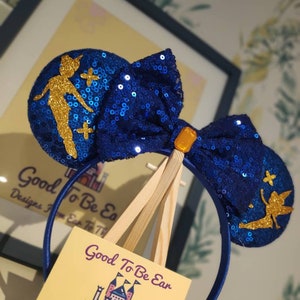 May include: Blue and gold sequined Minnie Mouse ears with a Tinkerbell silhouette on each ear. The headband is blue and the bow is blue with a gold center. The ears are displayed on a wooden easel with a business card that reads "Good To Be Ear, Designs From Ear To There".