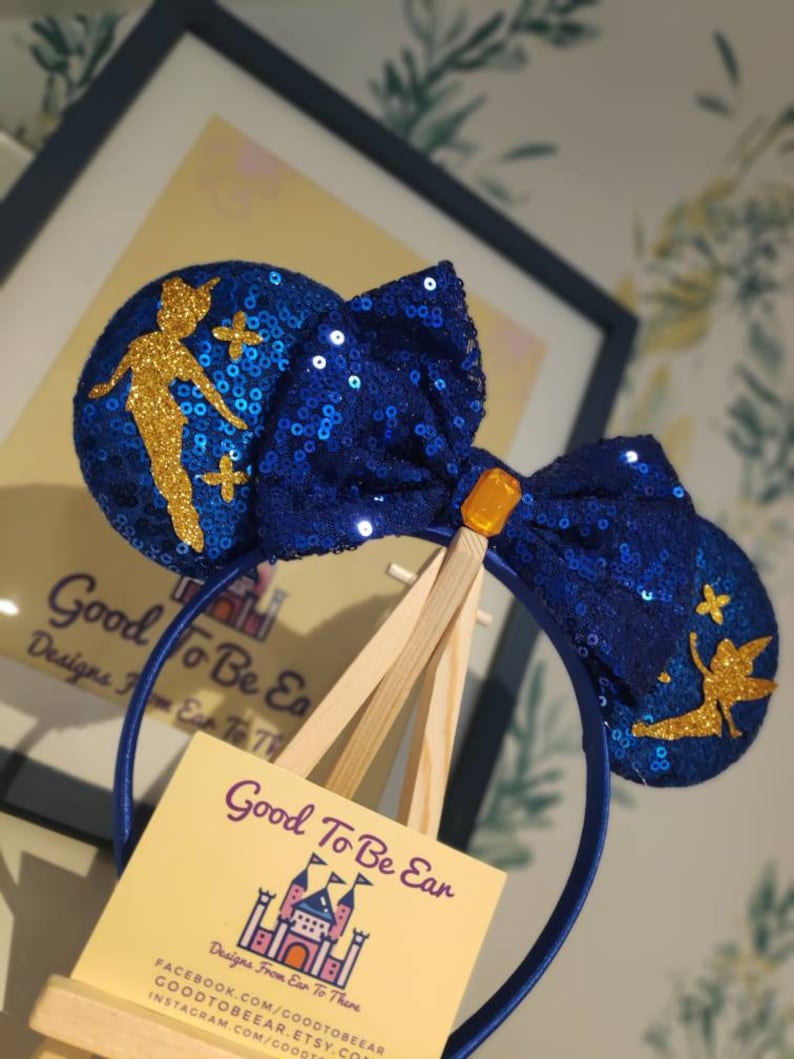 May include: Blue and gold sequined Minnie Mouse ears with a Tinkerbell silhouette on each ear. The headband is blue and the bow is blue with a gold center. The ears are displayed on a wooden easel with a business card that reads "Good To Be Ear, Designs From Ear To There".