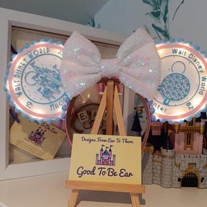May include: A white sequined bow displayed on a miniature wooden easel, with a sign reading "Good To Be Ear". Two illuminated Walt Disney World decorations are behind the bow. A small castle is visible in the background.