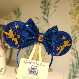 May include: Blue sequin Minnie Mouse ears with a gold bow and a Tinkerbell silhouette on each ear. The ears are on a wooden easel with a business card that reads "Good To Be Ear, Designs From Ear To There, Facebook.com/goodtobear, goodtobear.etsy.com, instagram.com/goodtobear".