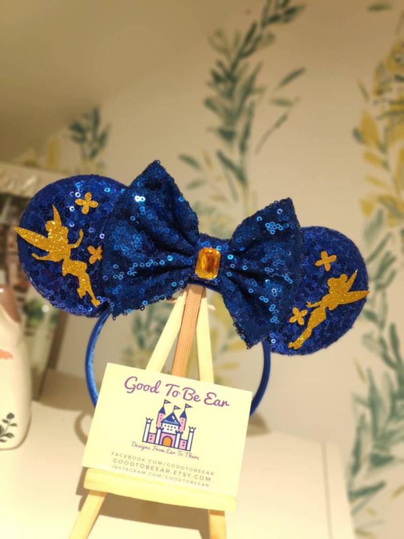 May include: Blue sequin Minnie Mouse ears with a gold bow and a Tinkerbell silhouette on each ear. The ears are on a wooden easel with a business card that reads "Good To Be Ear, Designs From Ear To There, Facebook.com/goodtobear, goodtobear.etsy.com, instagram.com/goodtobear".