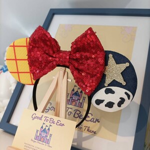 Op de afbeelding: Een rood pailletten strik hoofdband met een geel en rood geruit oor aan de ene kant en een blauw denim oor met een witte koe print en een gouden ster aan de andere kant. De hoofdband wordt getoond op een houten ezel met een visitekaartje waarop staat "Good To Be Ear Designs From Ear To There" met contactgegevens eronder.
