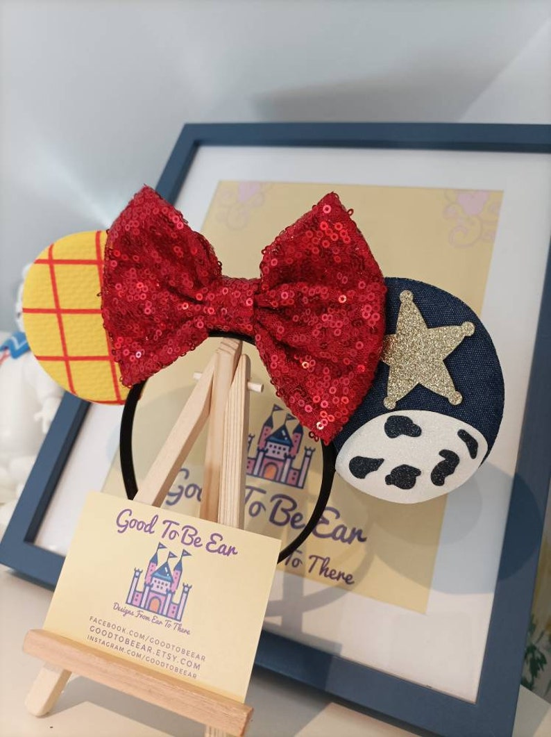 Op de afbeelding: Een rood pailletten strik hoofdband met een geel en rood geruit oor aan de ene kant en een blauw denim oor met een witte koe print en een gouden ster aan de andere kant. De hoofdband wordt getoond op een houten ezel met een visitekaartje waarop staat "Good To Be Ear Designs From Ear To There" met contactgegevens eronder.
