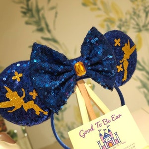 May include: Blue sequin Minnie Mouse ears with a gold Tinkerbell silhouette on each ear. The bow is blue with a gold gem in the center. The ears are on a wooden stand with a business card that reads "Good To Be Ear Designs From Ear To There" with contact information.