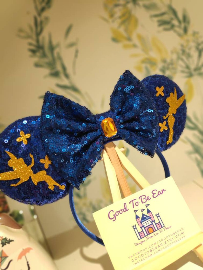 May include: Blue sequin Minnie Mouse ears with a gold Tinkerbell silhouette on each ear. The bow is blue with a gold gem in the center. The ears are on a wooden stand with a business card that reads "Good To Be Ear Designs From Ear To There" with contact information.