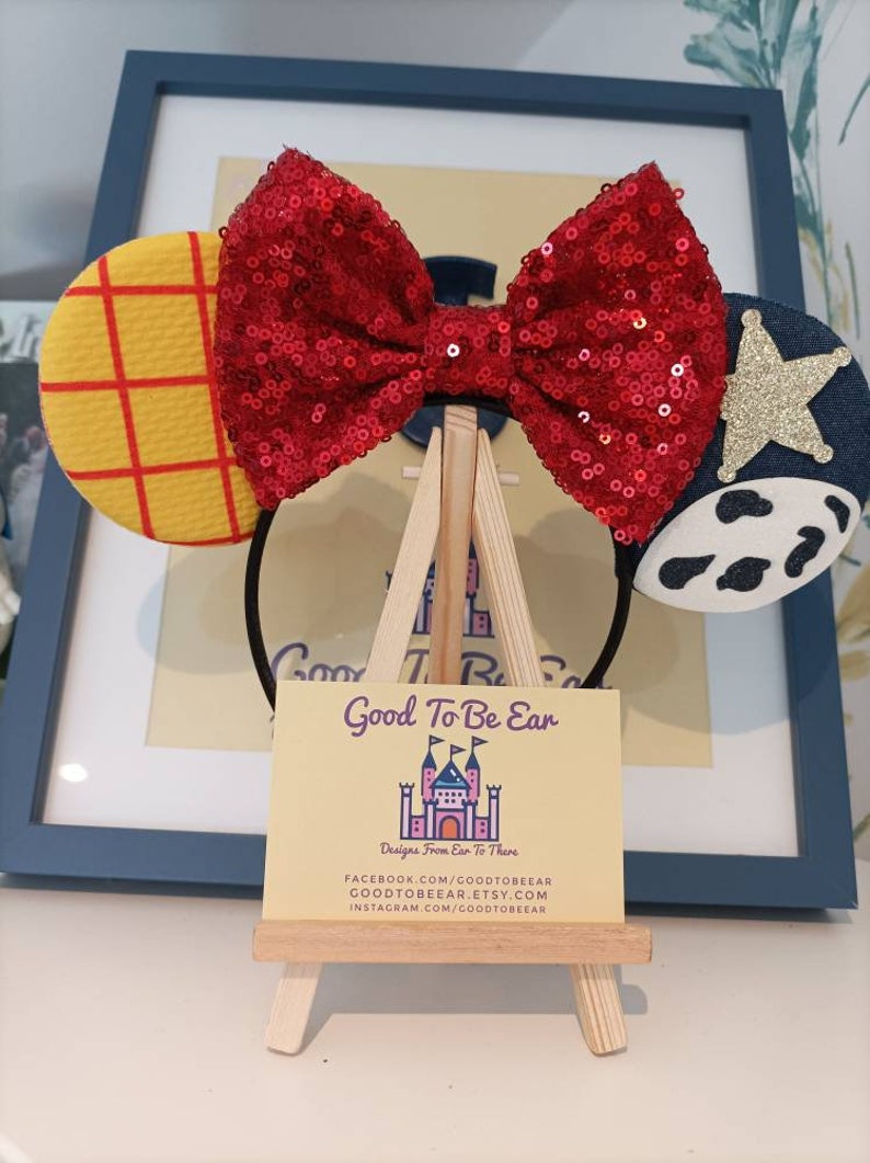 Op de afbeelding: Een rood pailletten strik hoofdband met een geel en rood geruit oor aan de ene kant en een blauw en wit koe-print oor aan de andere kant. Het hoofdband wordt getoond op een kleine houten ezel met een visitekaartje waarop staat "Good To Be Ear Designs From Ear To There" en sociale media handvatten.
