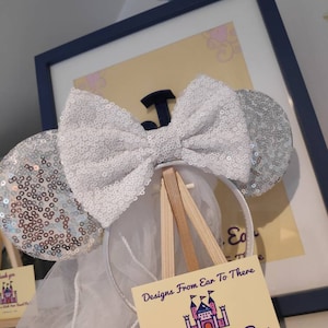 May include: White sparkly Minnie Mouse ears with a large white bow. The ears are on a wooden easel with a sign that reads "Designs From Ear To There Good To Be Ear".