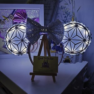 May include: Light up Minnie Mouse ears with a white bow and a geometric design. The ears are lit up with white lights and have a silver glitter finish. The ears are on a headband and are displayed on a small easel with a business card that reads "Good To Be Ear".