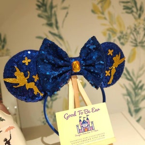 May include: Blue and gold sparkly Minnie Mouse ears with a Tinkerbell design. The ears have a blue bow with a gold gem in the center. The ears are on a wooden easel with a yellow sign that says "Good To Be Ear" and "Designs From Ear To There".