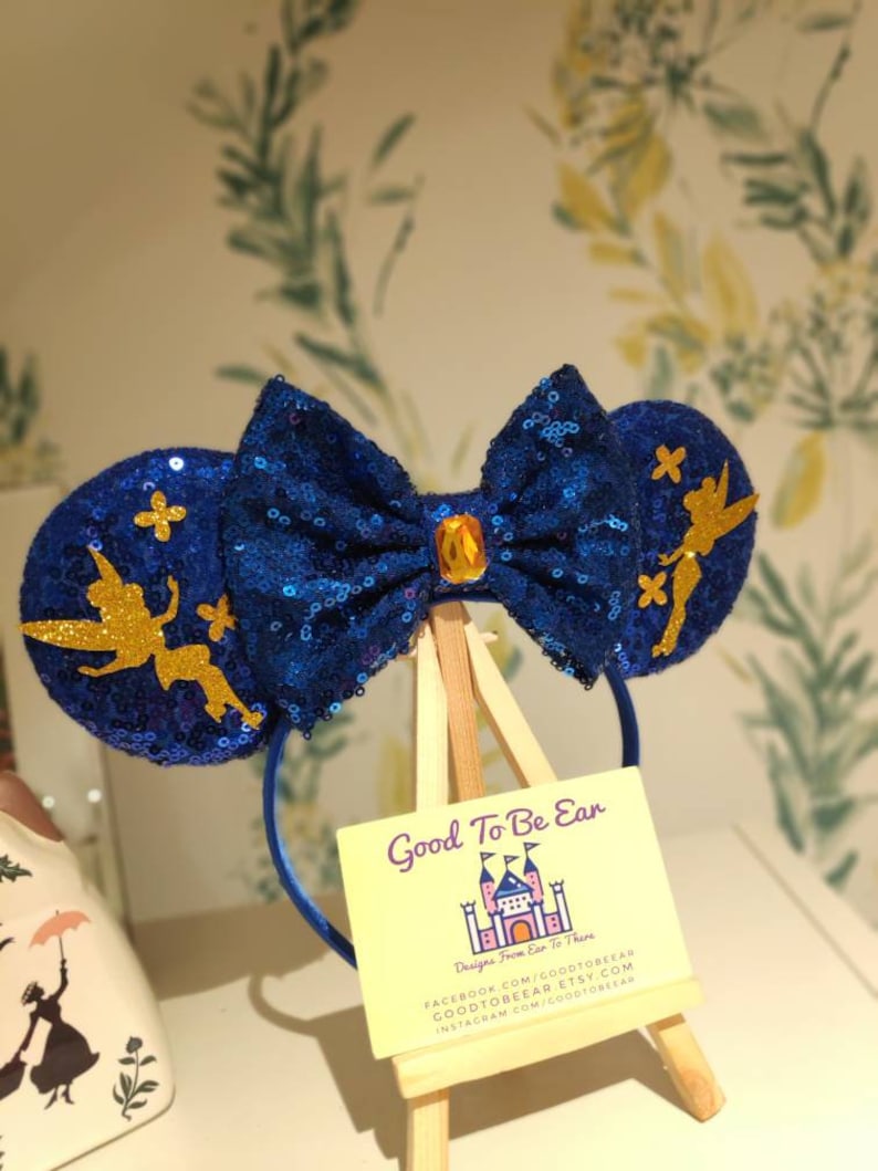 May include: Blue and gold sparkly Minnie Mouse ears with a Tinkerbell design. The ears have a blue bow with a gold gem in the center. The ears are on a wooden easel with a yellow sign that says "Good To Be Ear" and "Designs From Ear To There".