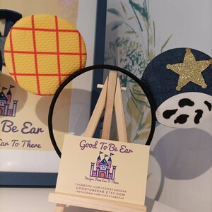 Op de afbeelding: Een paar handgemaakte Mickey Mouse oren met een geel en rood geruit patroon op het ene oor en een blauw denim oor met een gouden ster en witte koe vlekken op het andere. De oren worden getoond op een klein houten ezel met een visitekaartje waarop staat "Good To Be Ear Designs From Ear To There" en contactgegevens voor Facebook, Etsy en Instagram.