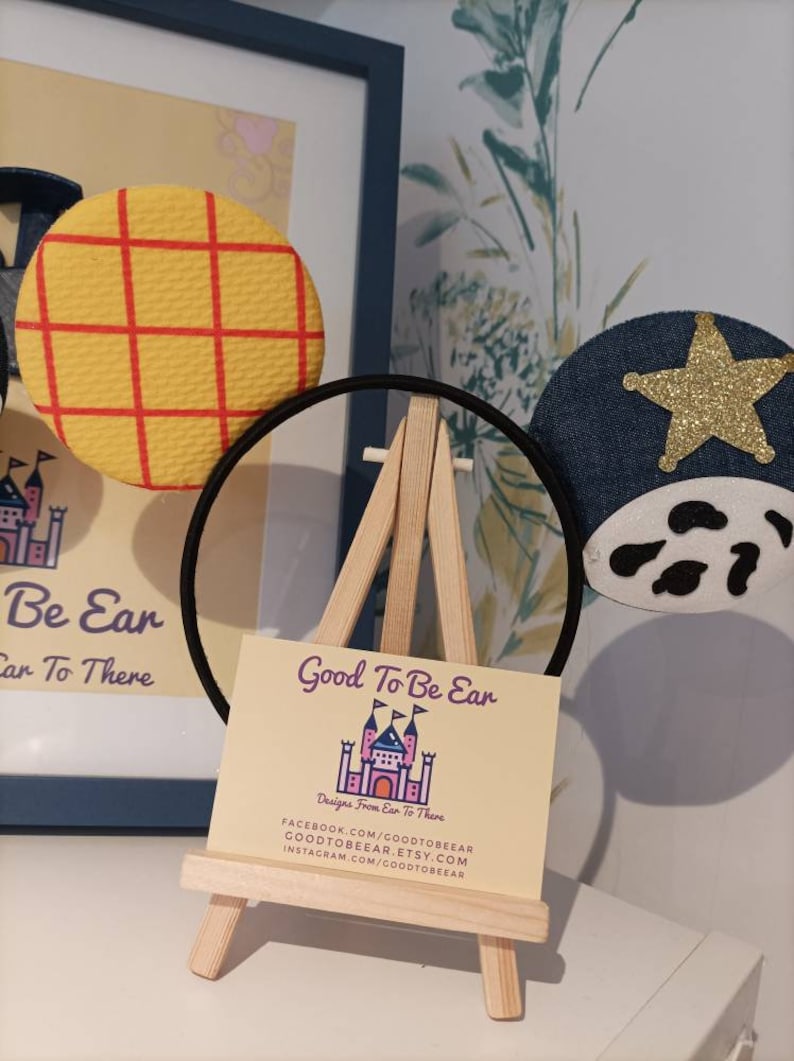 Op de afbeelding: Een paar handgemaakte Mickey Mouse oren met een geel en rood geruit patroon op het ene oor en een blauw denim oor met een gouden ster en witte koe vlekken op het andere. De oren worden getoond op een klein houten ezel met een visitekaartje waarop staat "Good To Be Ear Designs From Ear To There" en contactgegevens voor Facebook, Etsy en Instagram.