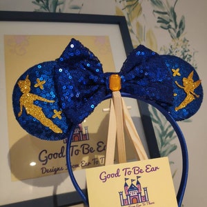 May include: Blue sequin Minnie Mouse ears with a gold Tinkerbell silhouette and a blue bow with a gold gem in the center. The ears are on a wooden easel with a business card that reads "Good To Be Ear Designs From Ear To There".