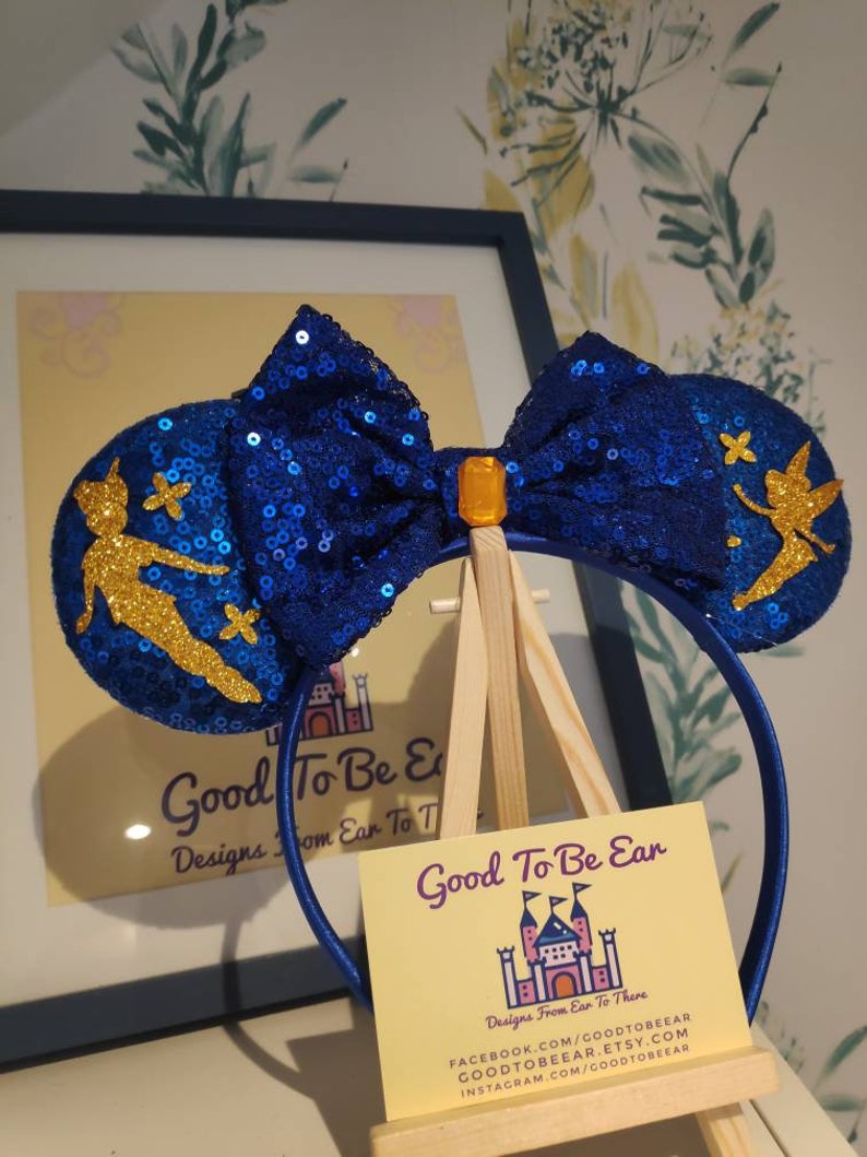 May include: Blue sequin Minnie Mouse ears with a gold Tinkerbell silhouette and a blue bow with a gold gem in the center. The ears are on a wooden easel with a business card that reads "Good To Be Ear Designs From Ear To There".