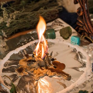 May include: A burning candle with melted wax on a silver surface surrounded by crystals and herbs. The candle is surrounded by a circle of white powder.