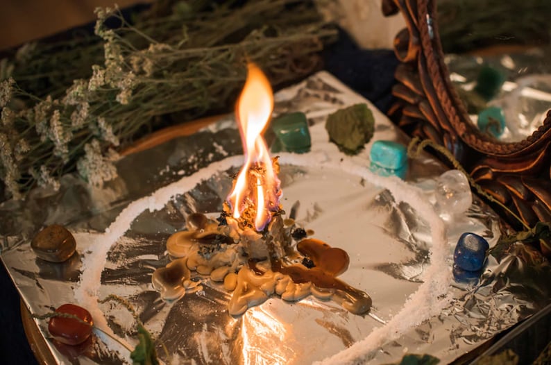 May include: A burning candle with melted wax on a silver surface surrounded by crystals and herbs. The candle is surrounded by a circle of white powder.