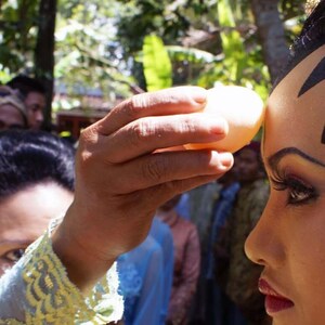 May include: A woman wearing a traditional headdress with gold and white embellishments has a white egg placed on her forehead during a ceremony.