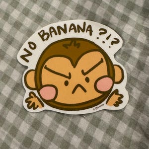 May include: A die-cut sticker featuring a brown monkey with a tan face, pink cheeks, and an angry expression. The text NO BANANA ?!? is printed in black capital letters in an arc above the monkey's head. The sticker has a white border.
