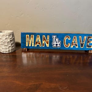 May include: A blue wooden sign with the words "MAN LA CAVE" in a burnt wood style. The sign has a Los Angeles Dodgers logo. A textured, light-colored vase sits to the left of the sign.