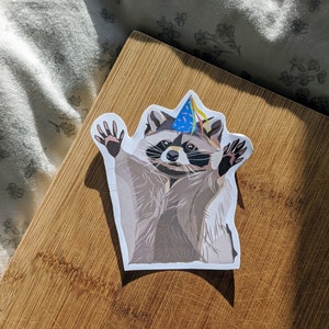 Könnte beinhalten: Ein gestanzter Aufkleber mit einem Waschbären, der einen Partyhut mit Regenbogen-Luftschlangen trägt. Der Waschbär hat die Arme erhoben. Der Aufkleber befindet sich auf einer Holzoberfläche.