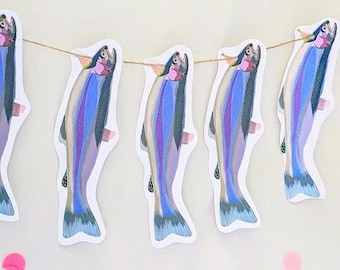 May include: A string of paper cut-out fish in shades of blue, purple, and gray, strung on a thin twine. The fish are arranged in a row, with pink confetti scattered around. The fish have a whimsical, artistic design.