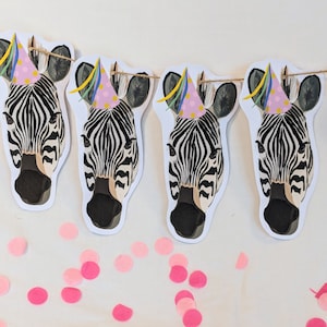 Puede incluir: Una guirnalda festiva con cuatro cabezas de cebra adornadas con sombreros de fiesta rosas y serpentinas de colores. Las cebras tienen rayas blancas y negras. Confeti rosa esparcido debajo.