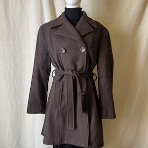May include: A dark brown double-breasted coat featuring a wide collar, long sleeves, and a matching fabric tie belt at the waist. The coat includes two front pockets and is worn over a black turtleneck sweater, displaying a structured silhouette.