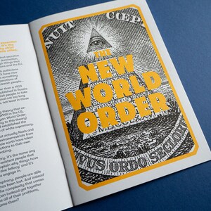May include: A black and white illustration of a pyramid with an eye at the top. The pyramid is surrounded by a yellow border with the text "The New World Order" in large yellow letters. The text "Novus Ordo Seclorum" is printed in black at the bottom of the image.