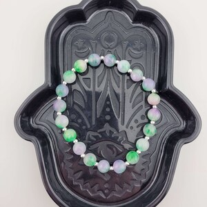 May include: A green and purple gemstone bracelet with silver beads, displayed on a black hamsa hand dish.