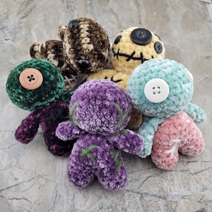 May include: Five crocheted plush toys in various colors and textures. The toys are all different shapes and sizes, and they have different facial expressions. The toys are arranged in a circle on a gray surface.