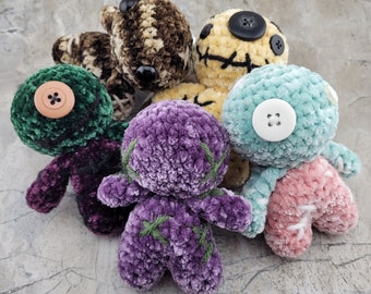 May include: Five crocheted plush toys in various colors and textures. The toys are all different shapes and sizes, and they have different facial expressions. The toys are arranged in a circle on a gray surface.