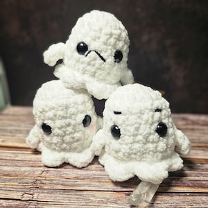 May include: Three white crocheted ghost plushies with black eyes and frowning expressions. The ghosts are stacked on top of each other.