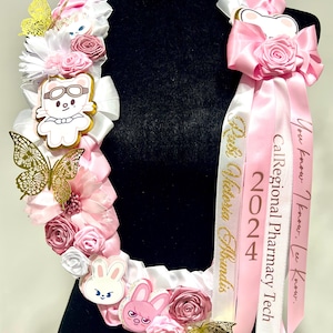 May include: A pink and white lei with gold butterflies, flowers, and cartoon animal charms. The lei has a pink ribbon with the text "CalRegional Pharmacy Tech 2024 Rubi Victoria Abundis" in gold lettering.